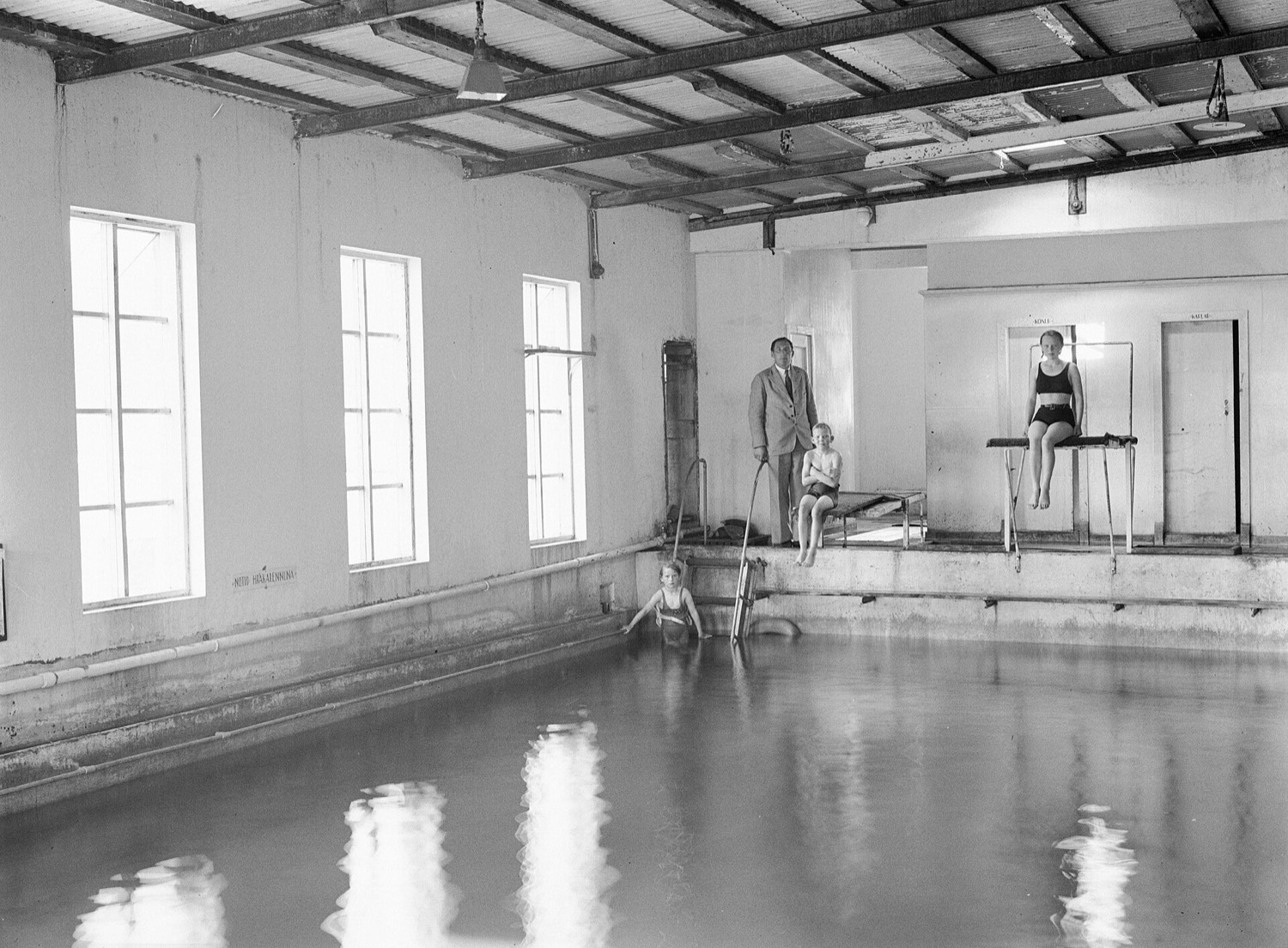 Bare interior of an indoor pool with a sheet metal roof: a man in a suit and tie holds a long pole at the edge, a child is in the water at the ladder, two girls sitting on the diving board