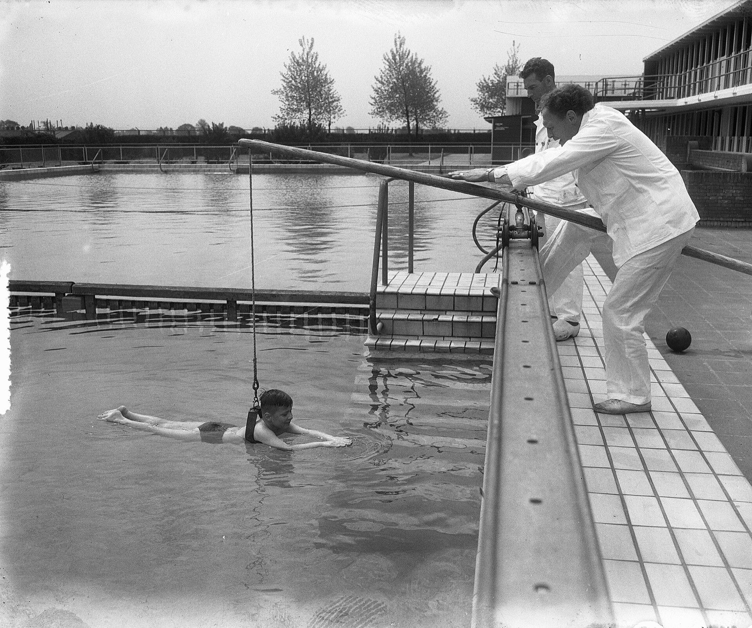 A child supported by a harness hanging from a sliding rope swims in the outdoor pool while two instructors in white slide the pole from the edge