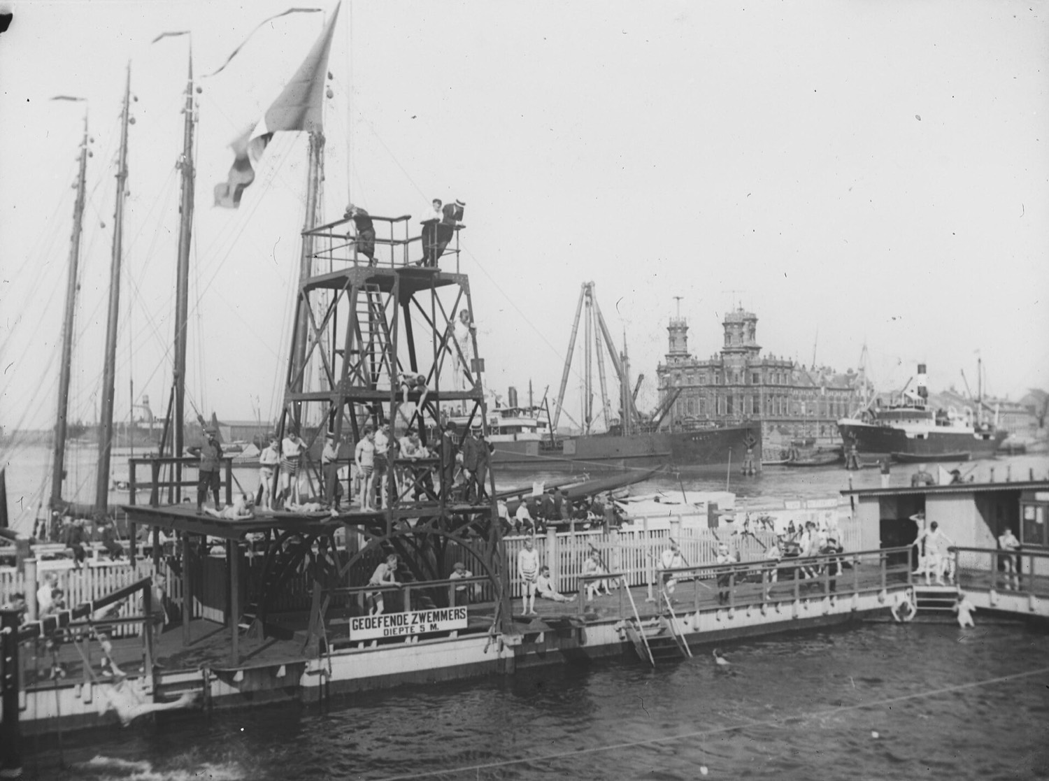 A wooden diving tower covered in boys at a floating establishment, with dozens of bodies on the piers and in the water