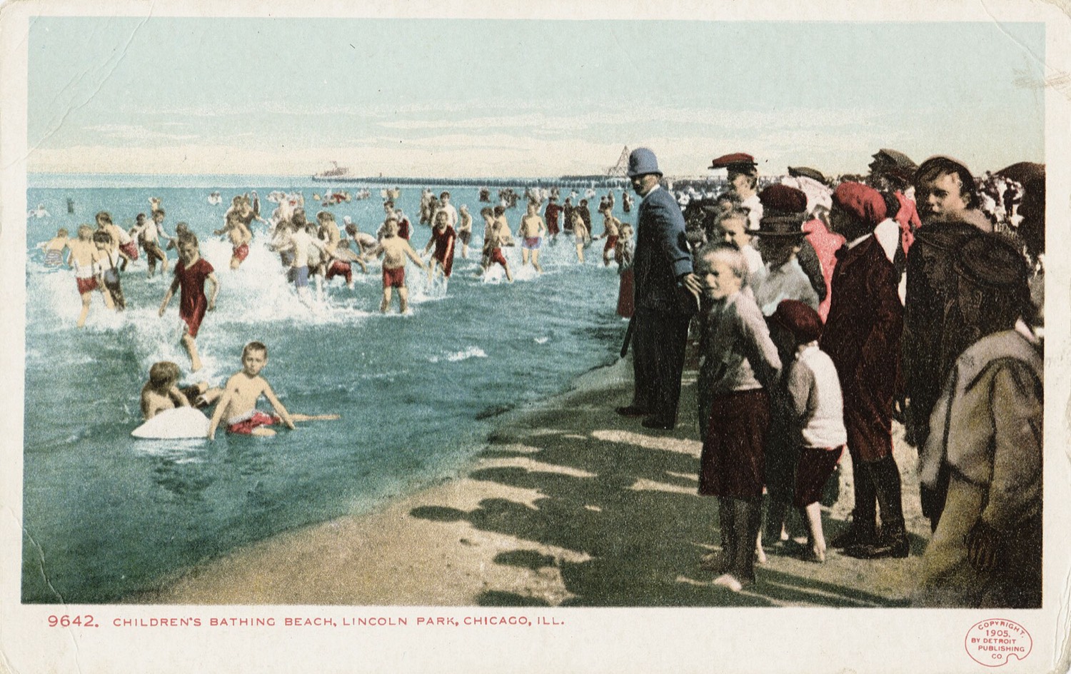 Dozens of children run into the water from an urban beach while a policeman holds back the crowd dressed on the shore