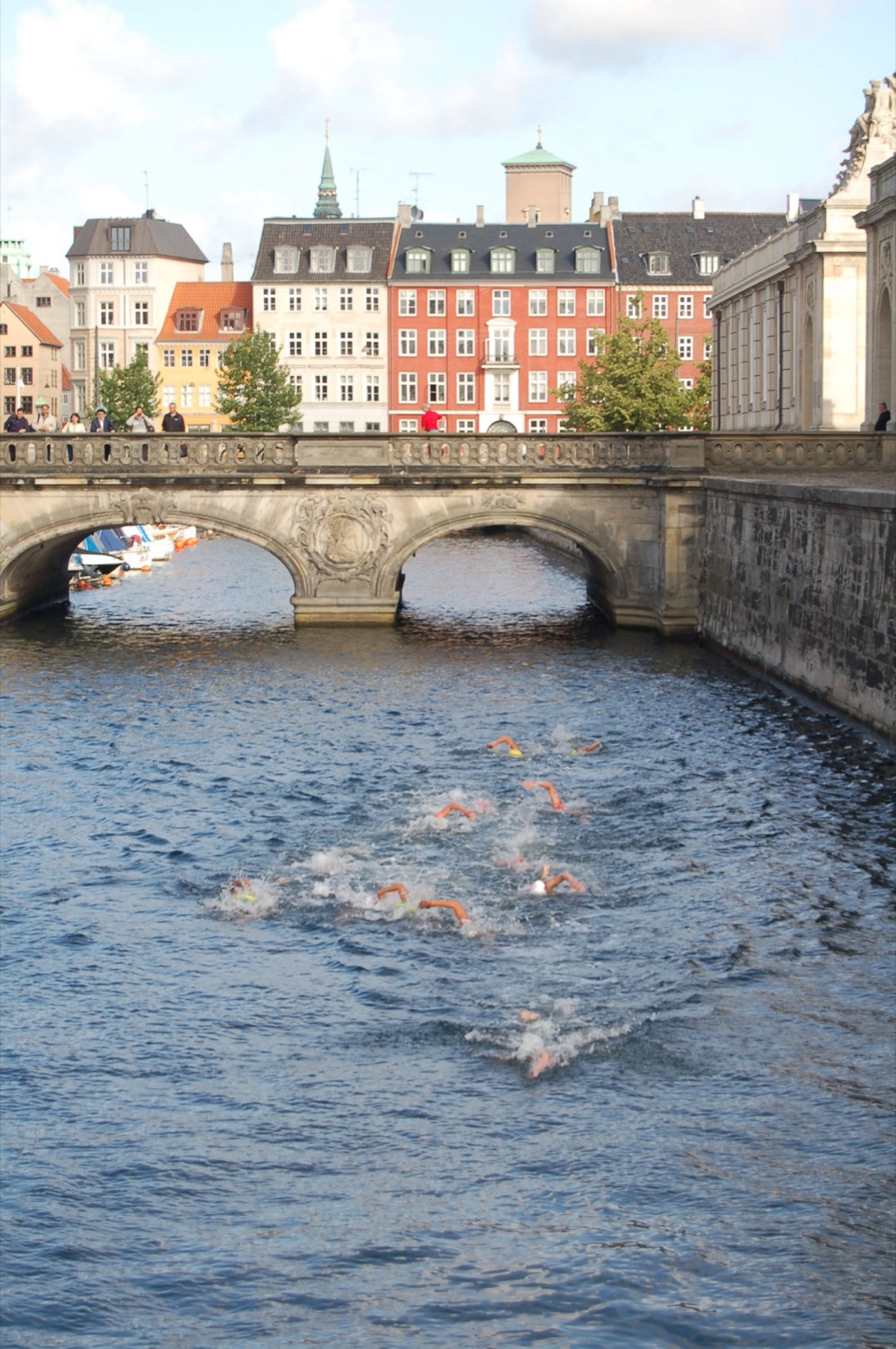 Swimmers with orange caps lined up in a canal under a carved stone arch, among red and yellow houses
