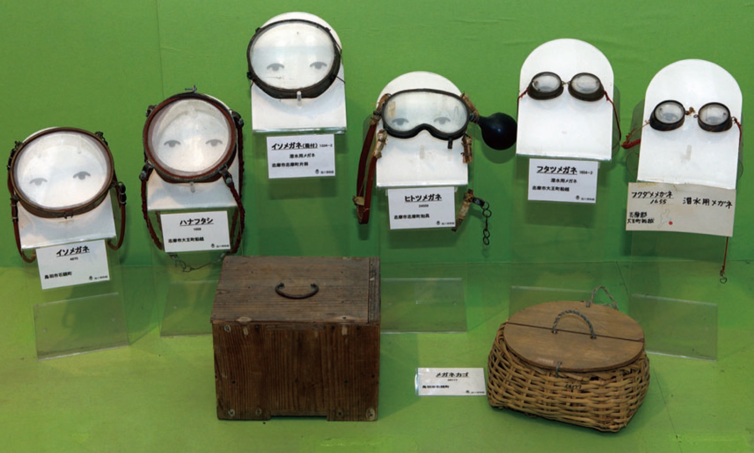 Six pairs of diving goggles displayed in a row on a green background, from the large single-lens with a leather frame to the two small glass portholes; the fourth one has a black rubber bag on the side