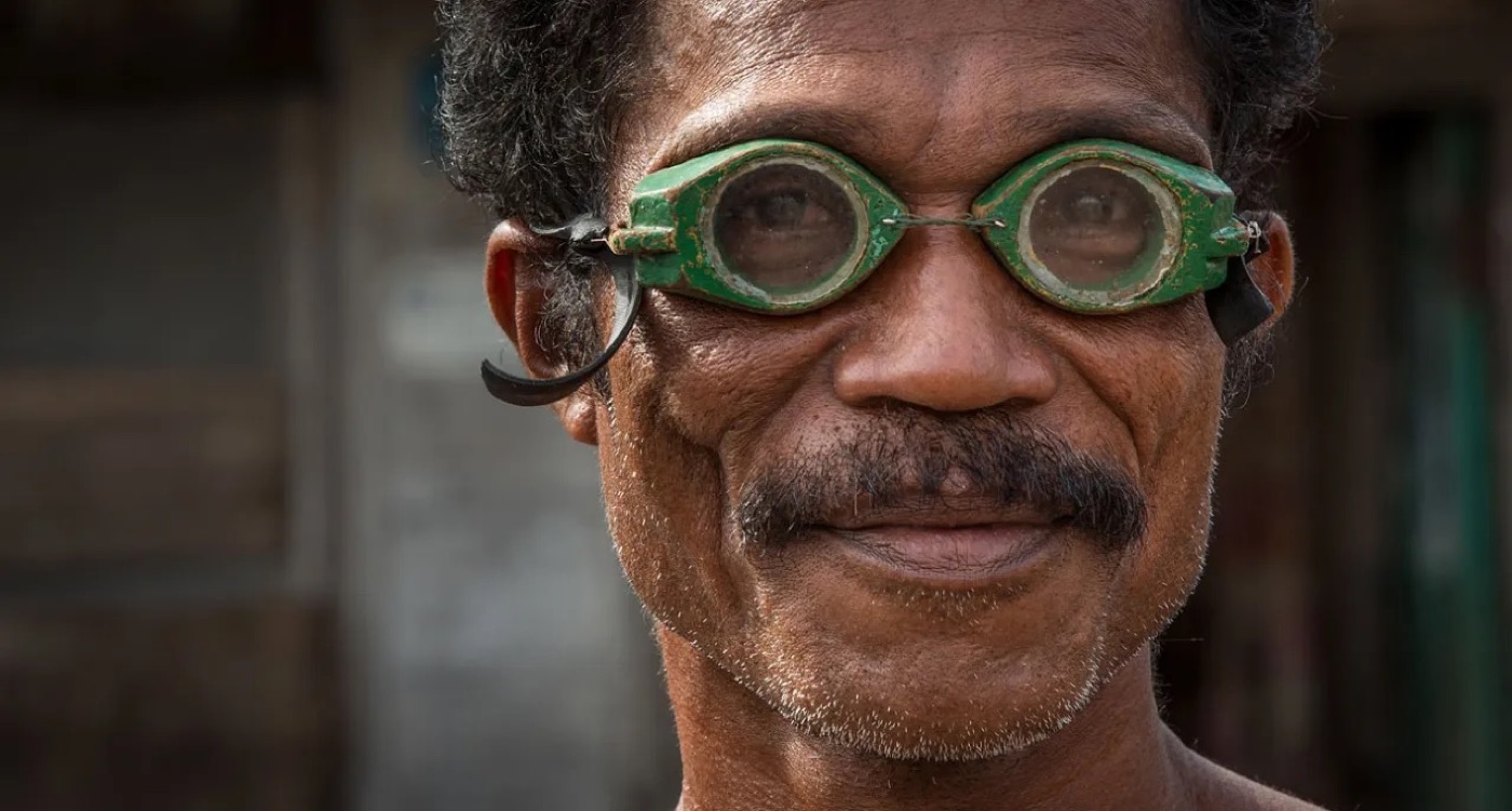 Close-up of a Bajau fisherman wearing hand-carved wooden goggles painted green, with two round glass lenses held by a metal wire and a rubber strap behind the ears