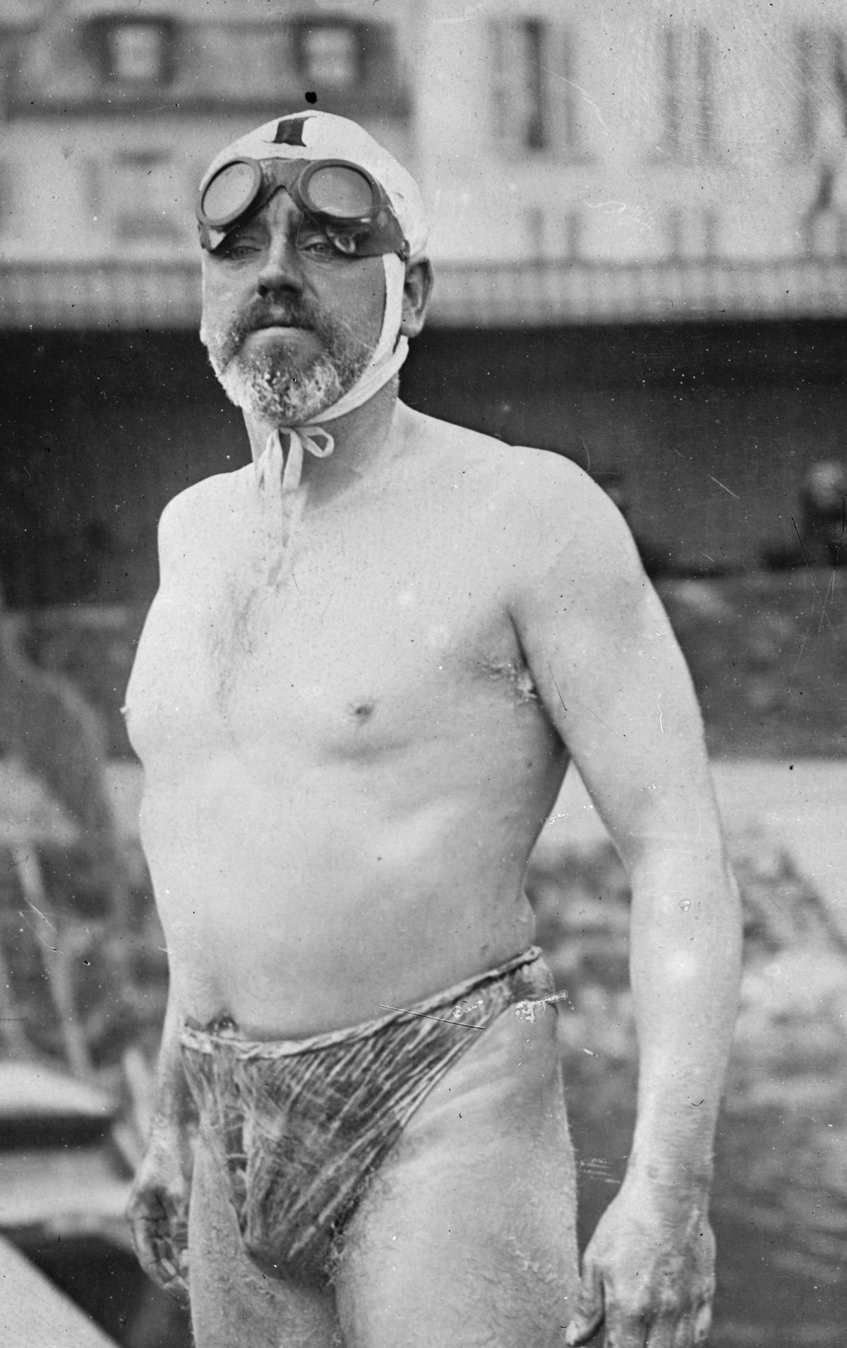 Bill Burgess standing on a pier in 1911, his body covered in grease, wearing a wool swimsuit and motorcycle goggles pushed up onto his forehead over his swim cap tied under his chin