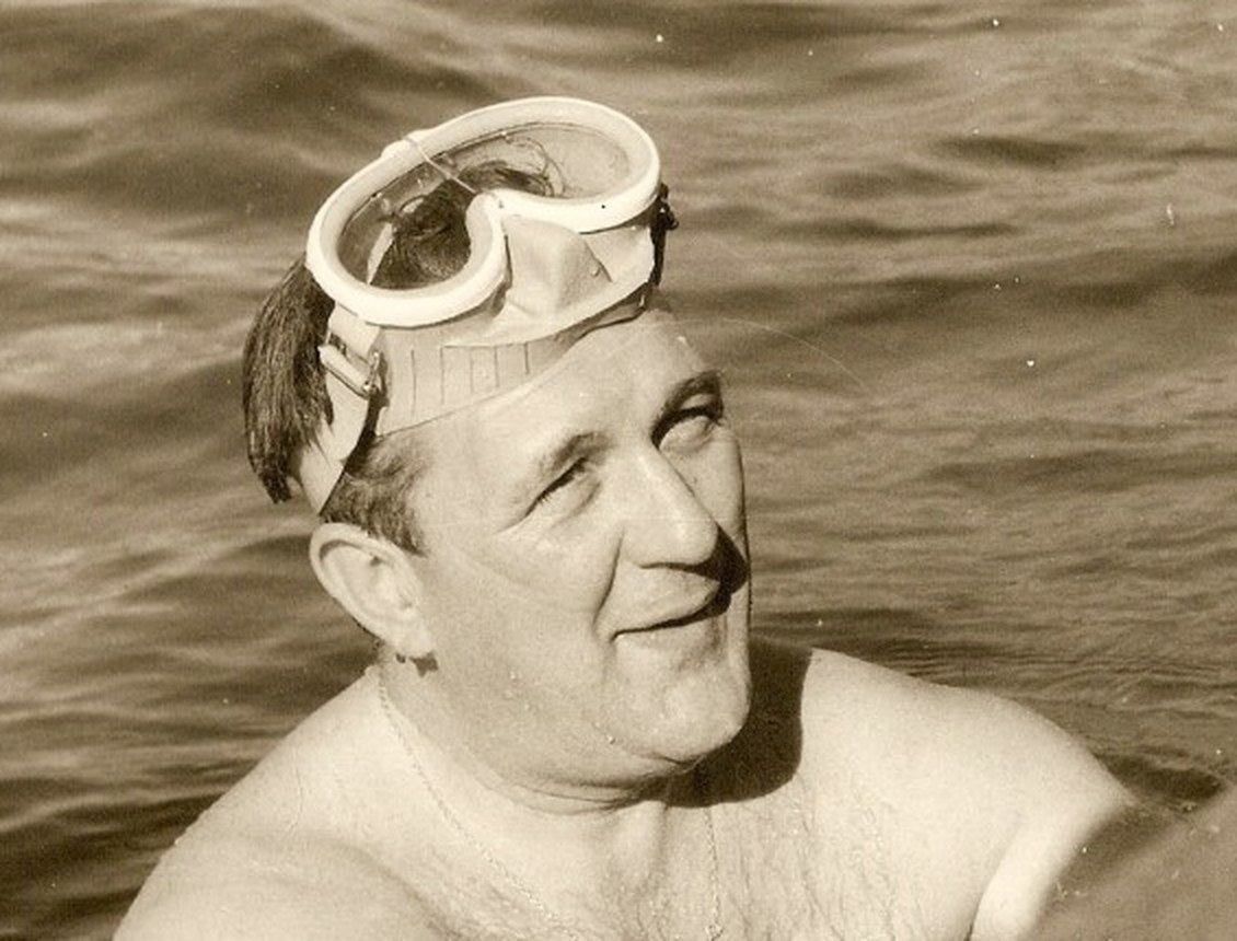 Luigi Ferraro surfaces from the water smiling, with the Pinocchio mask pushed onto his forehead: light frame, single lens, and the rubber nose pocket clearly visible