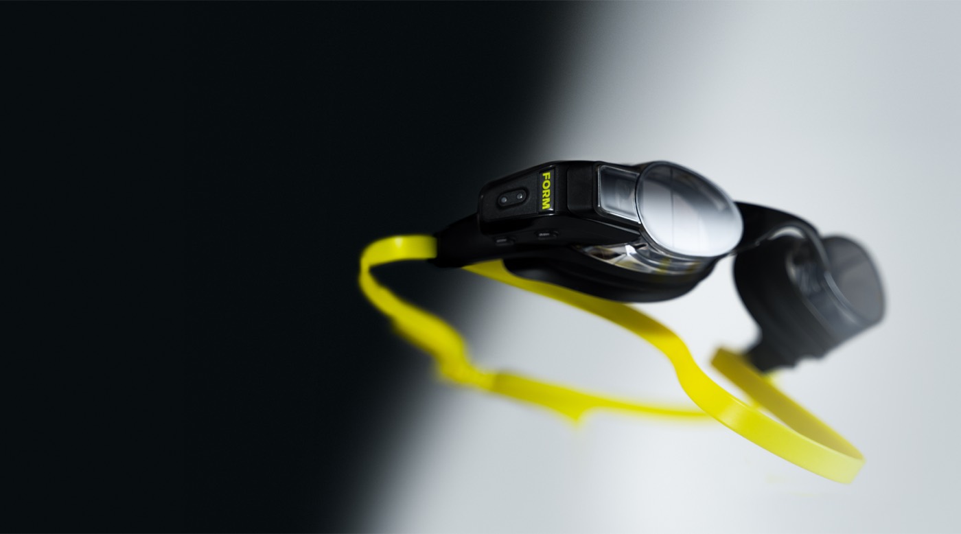 A pair of FORM Smart Swim goggles on a black and white background: the electronics module protrudes from the side of the right lens, the strap is yellow
