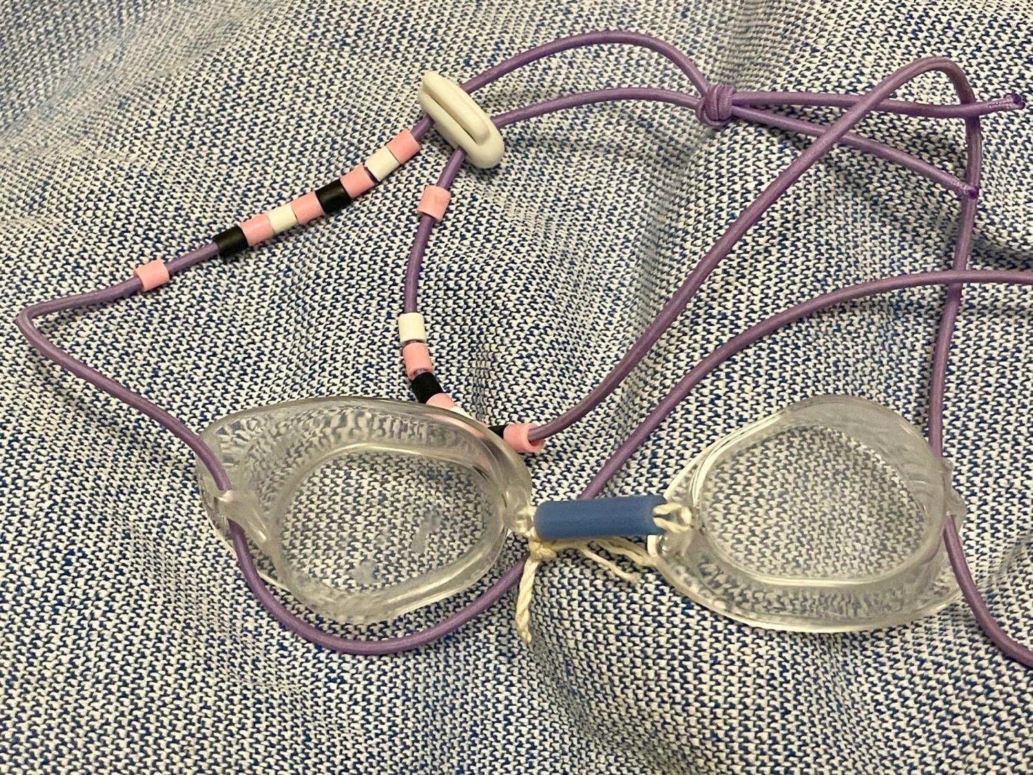A pair of Swedish goggles assembled by hand on a towel: two bare transparent lenses, the purple string with the spacers threaded one by one, a rubber tube, and a string tied in place of the nose bridge