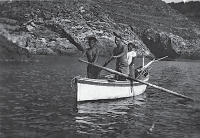 Black and white photograph: three men standing on a white gozzo moored under a rocky coast, with long bamboo poles resting across the boat