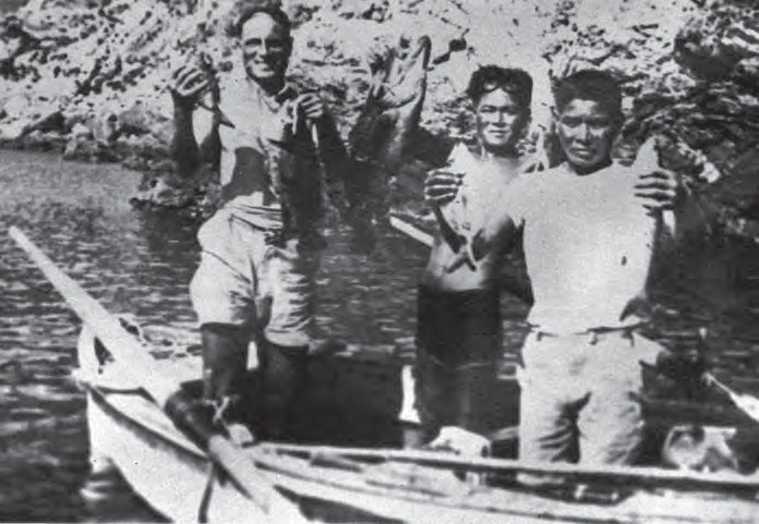Black and white photograph: three men standing on a boat lifting freshly caught groupers; the one in the center has his wooden goggles raised on his forehead