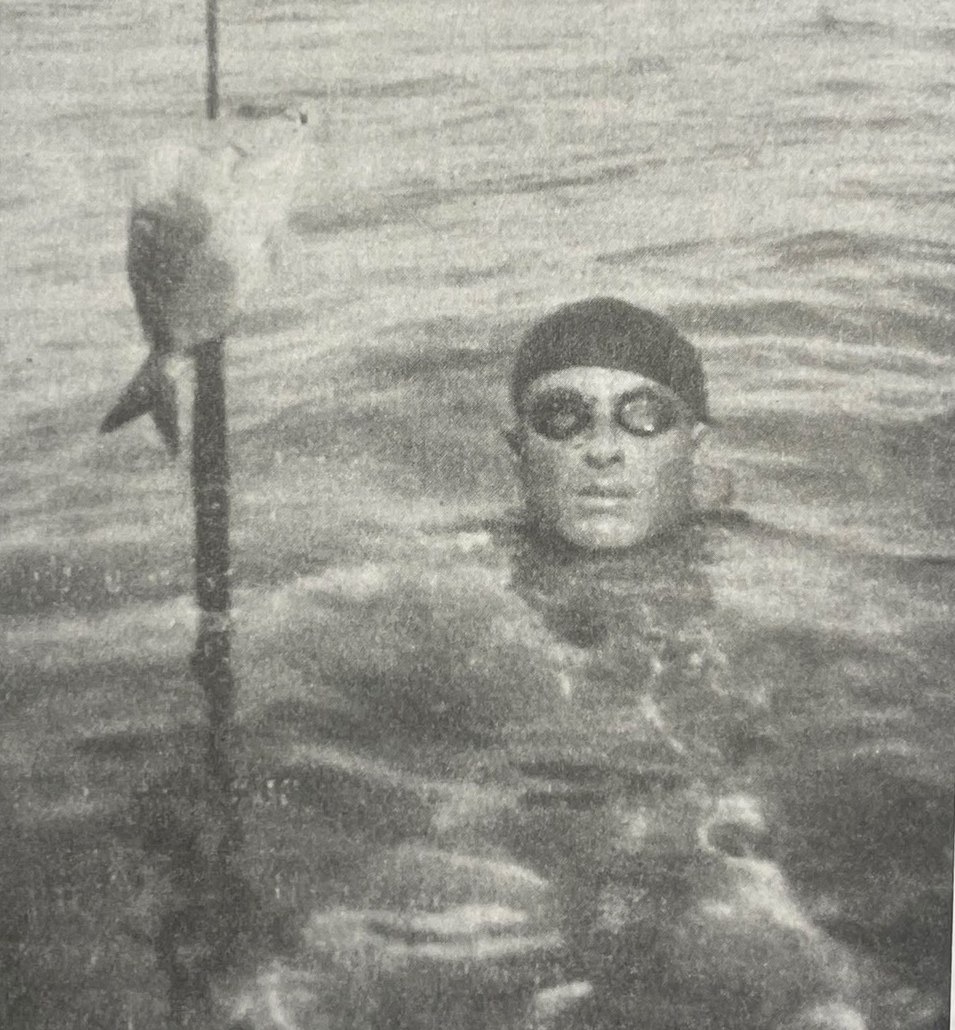 Black and white photograph: a man with a dark swim cap and round goggles emerges from the water next to a spear with a speared fish