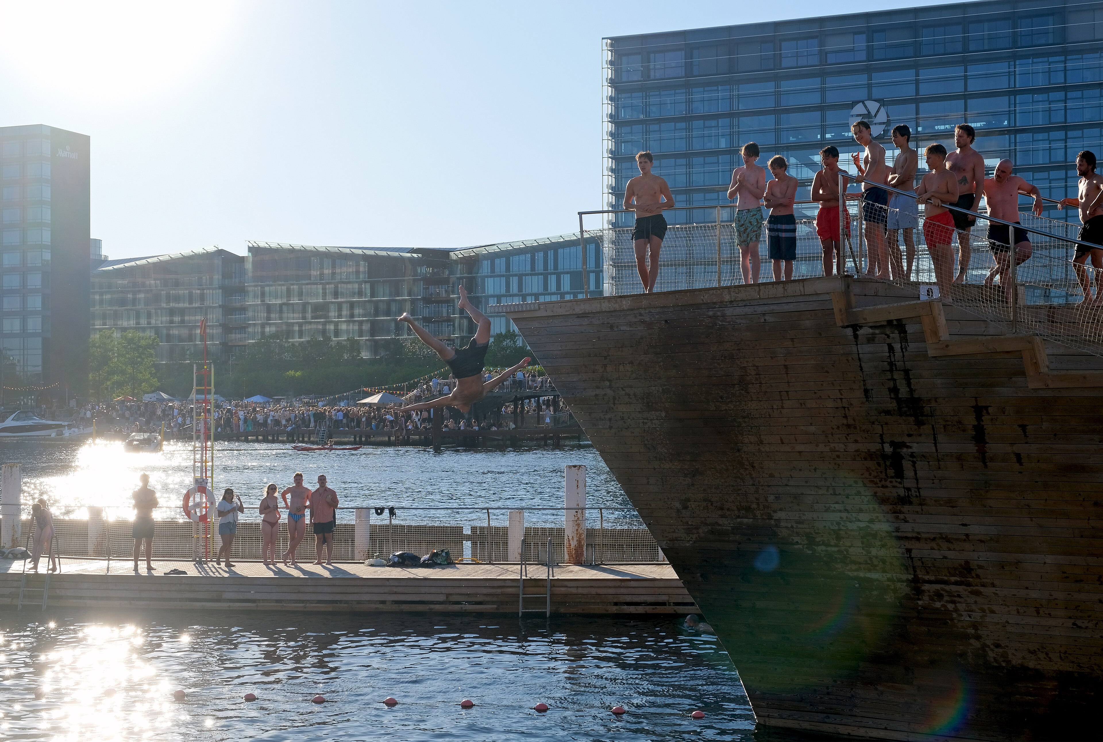 A group of people dives from the pier of the Islands Brygge harbour bath in Copenhagen, with the dark green water of the canal and the city in the background