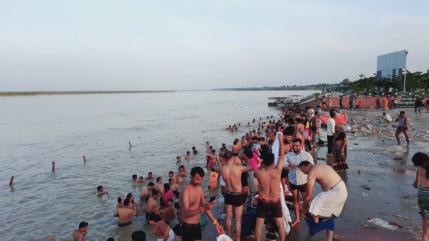 Hundreds of people in the water on the banks of the Ganges on a feast day, with waste accumulated on the sand in the foreground